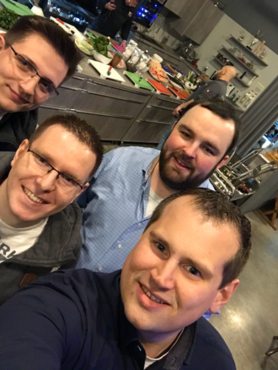 Gruppe von vier Männern in einer modernen Küche Vier Männer, die in einer modernen Küche lächeln, während im Hintergrund Essen zubereitet wird.